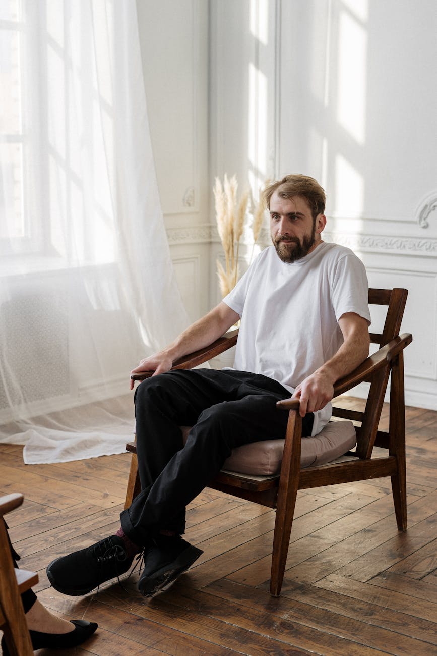 man in white crew neck t shirt sitting on brown wooden armchair